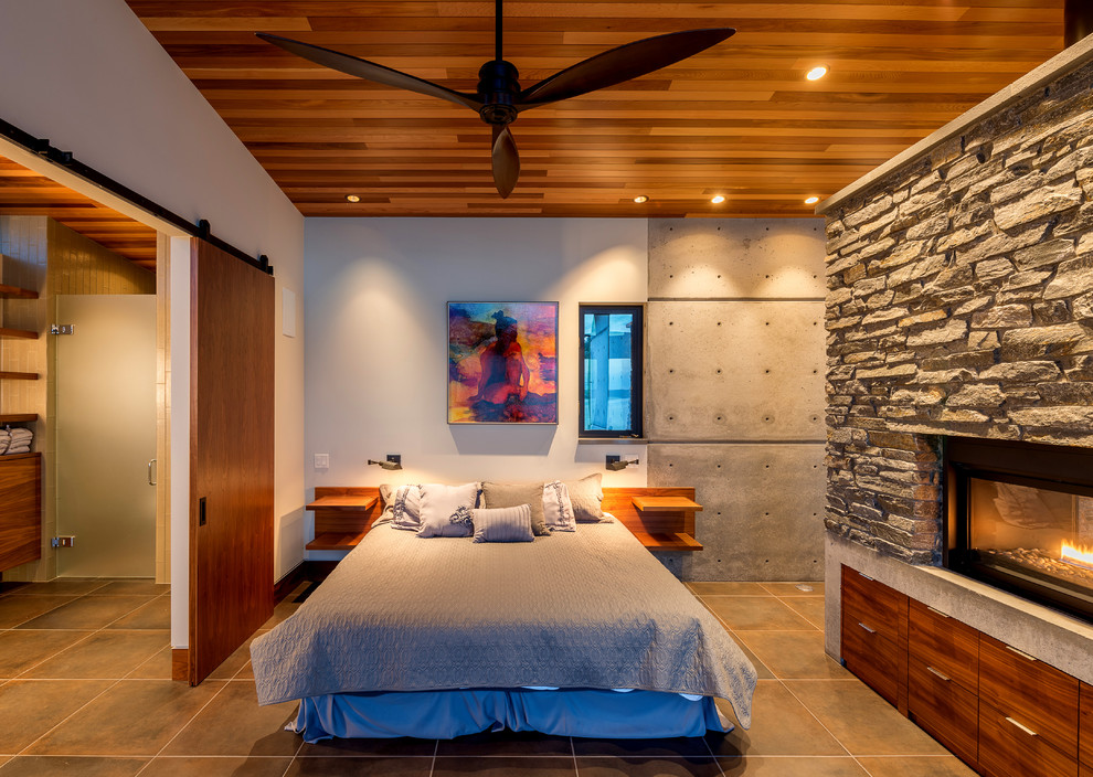 Imagen de dormitorio tipo loft moderno pequeño con paredes blancas, suelo de baldosas de porcelana, chimenea de doble cara, marco de chimenea de piedra y suelo gris