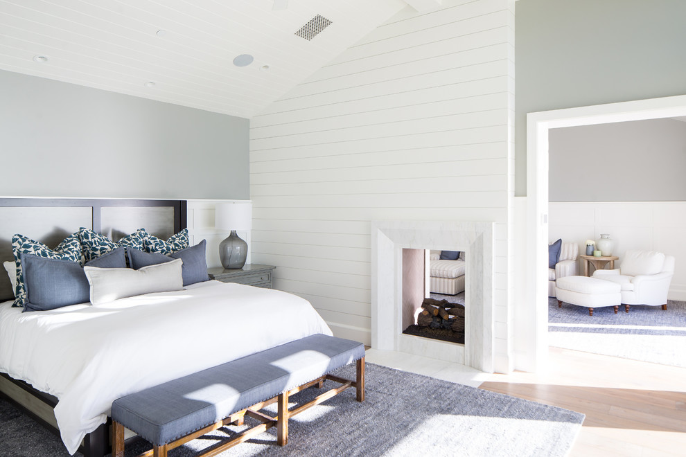 Modelo de dormitorio costero con paredes grises, suelo de madera clara, chimenea de doble cara y marco de chimenea de madera