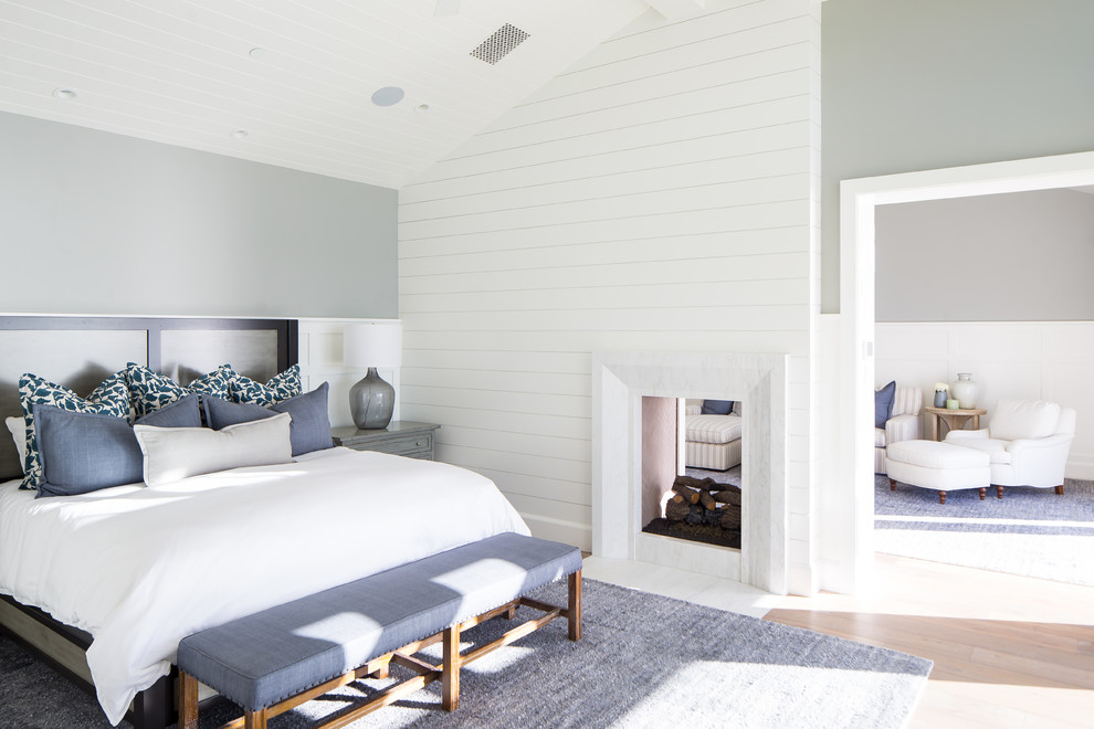 Modelo de dormitorio principal marinero con paredes grises, suelo de madera clara, chimenea de doble cara, marco de chimenea de piedra y suelo beige