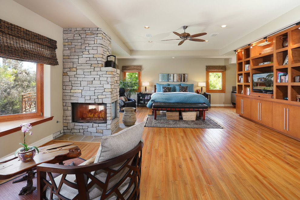 Elegant medium tone wood floor bedroom photo in Orange County with beige walls, a two-sided fireplace and a stone fireplace