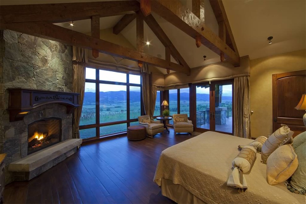 Foto de dormitorio principal rural grande con paredes beige, suelo de madera en tonos medios, chimenea de esquina y marco de chimenea de piedra