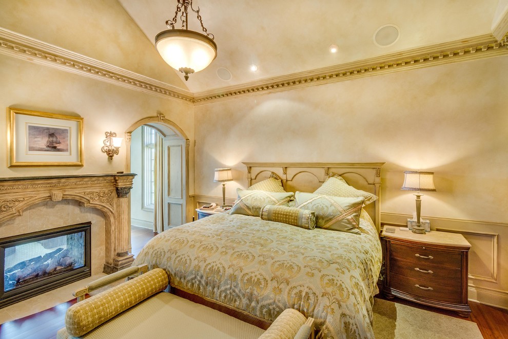 Imagen de dormitorio principal clásico grande con paredes beige, suelo de madera en tonos medios, chimenea de doble cara, marco de chimenea de yeso y suelo marrón