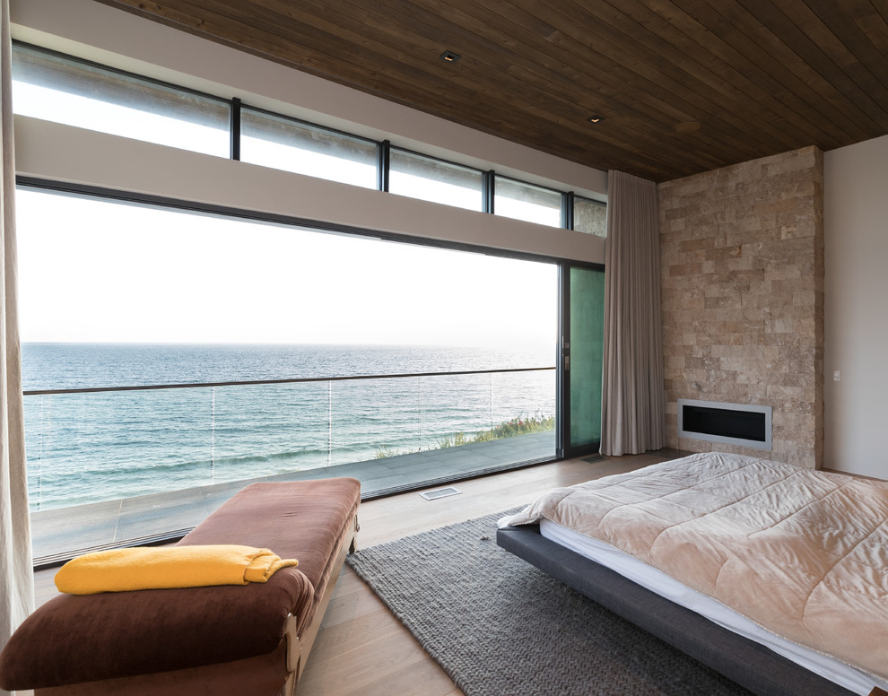 Imagen de dormitorio principal contemporáneo grande con paredes blancas, suelo de madera clara, chimenea lineal, marco de chimenea de piedra y suelo marrón