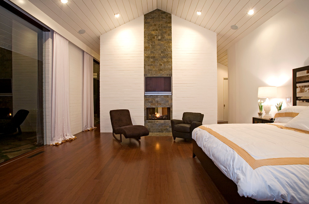 Imagen de dormitorio retro con paredes blancas, suelo de madera oscura, marco de chimenea de piedra y chimenea de doble cara