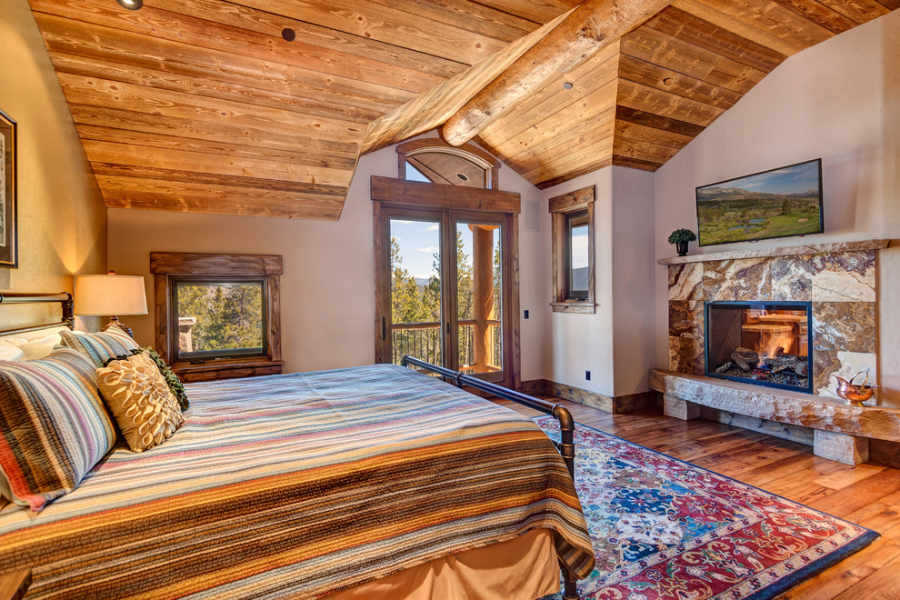 Diseño de dormitorio principal rústico grande con paredes beige, suelo de madera en tonos medios, todas las chimeneas, marco de chimenea de piedra y suelo marrón