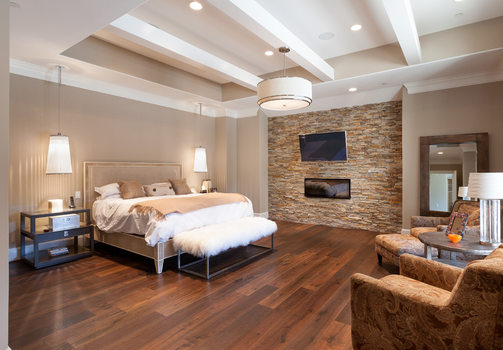 Modelo de dormitorio principal y televisión clásico renovado grande con chimenea lineal, paredes beige, suelo de madera oscura y marco de chimenea de piedra