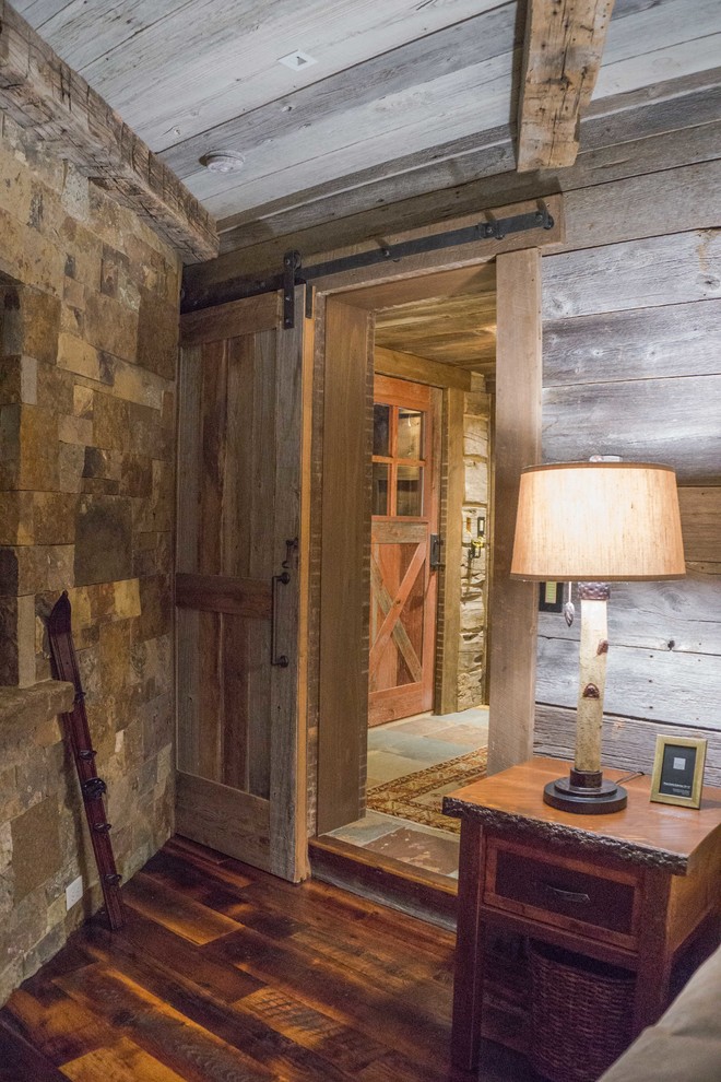 Imagen de dormitorio principal rural con suelo de madera en tonos medios, chimenea de esquina y marco de chimenea de piedra