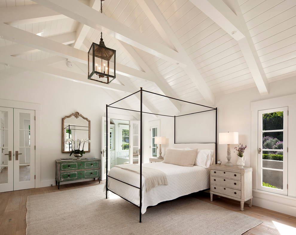 Farmhouse master bedroom in Santa Barbara with white walls, light hardwood flooring and feature lighting.