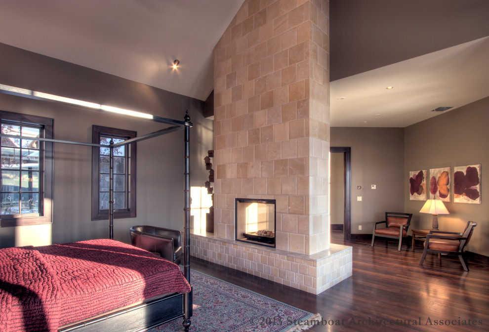 Cette photo montre une très grande chambre parentale moderne avec un sol en bois brun, une cheminée double-face et un manteau de cheminée en carrelage.