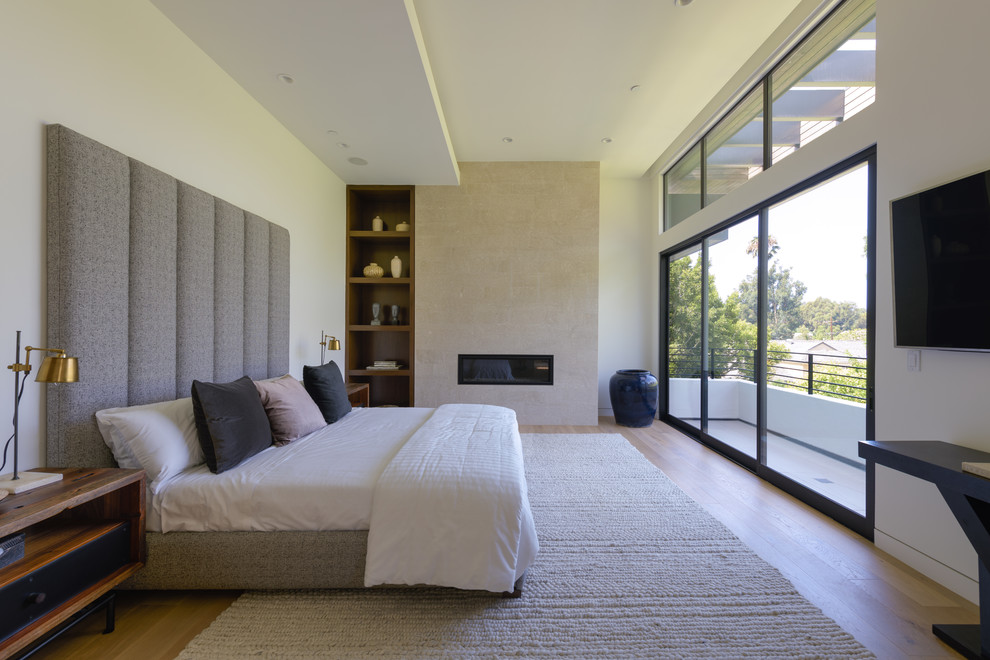Modelo de dormitorio moderno con paredes blancas, suelo de madera en tonos medios, chimenea lineal, marco de chimenea de baldosas y/o azulejos y suelo marrón