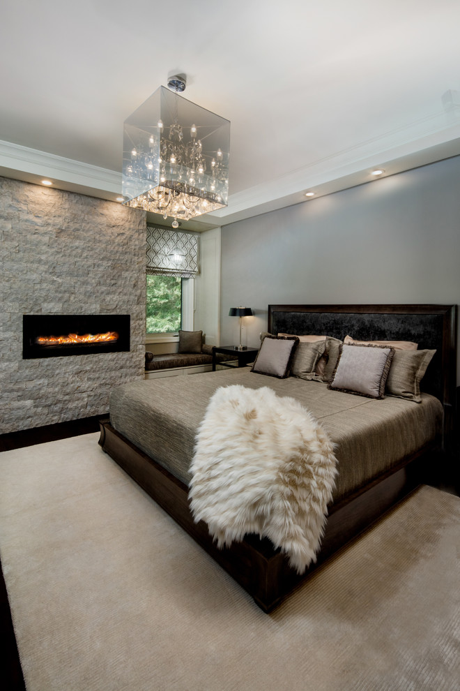 Cette image montre une grande chambre parentale design avec un mur gris, parquet foncé, une cheminée standard, un manteau de cheminée en pierre et un sol marron.