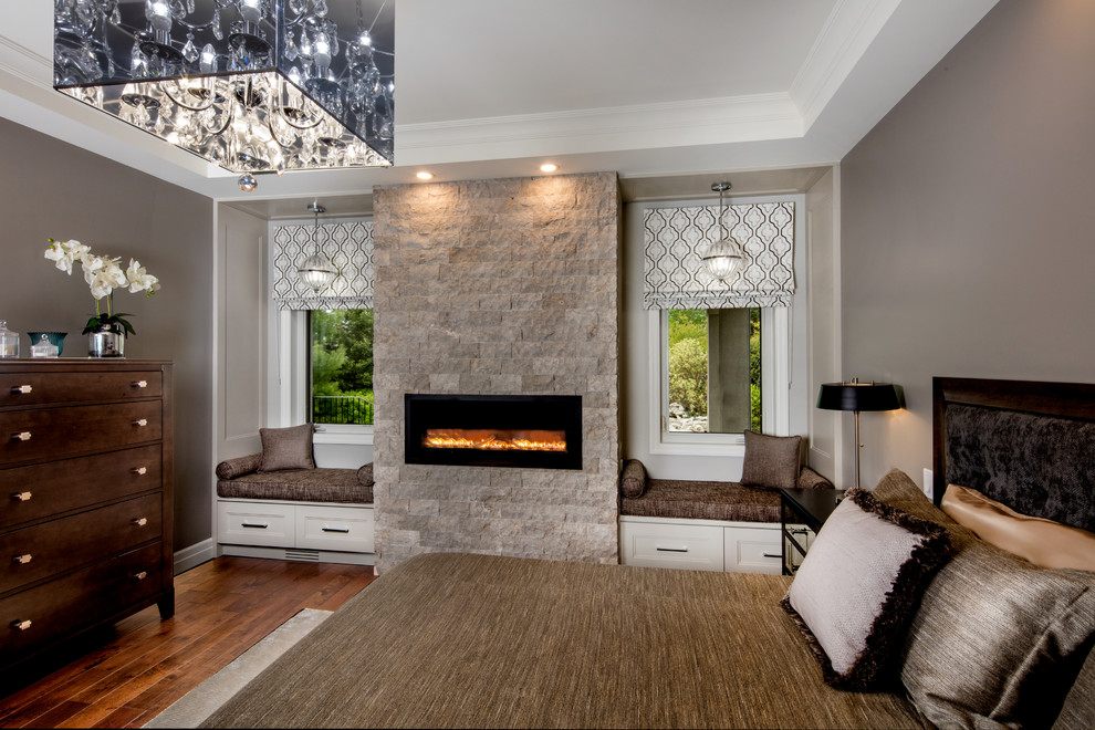 Cette image montre une grande chambre parentale design avec un mur gris, parquet foncé, une cheminée standard, un manteau de cheminée en pierre et un sol marron.