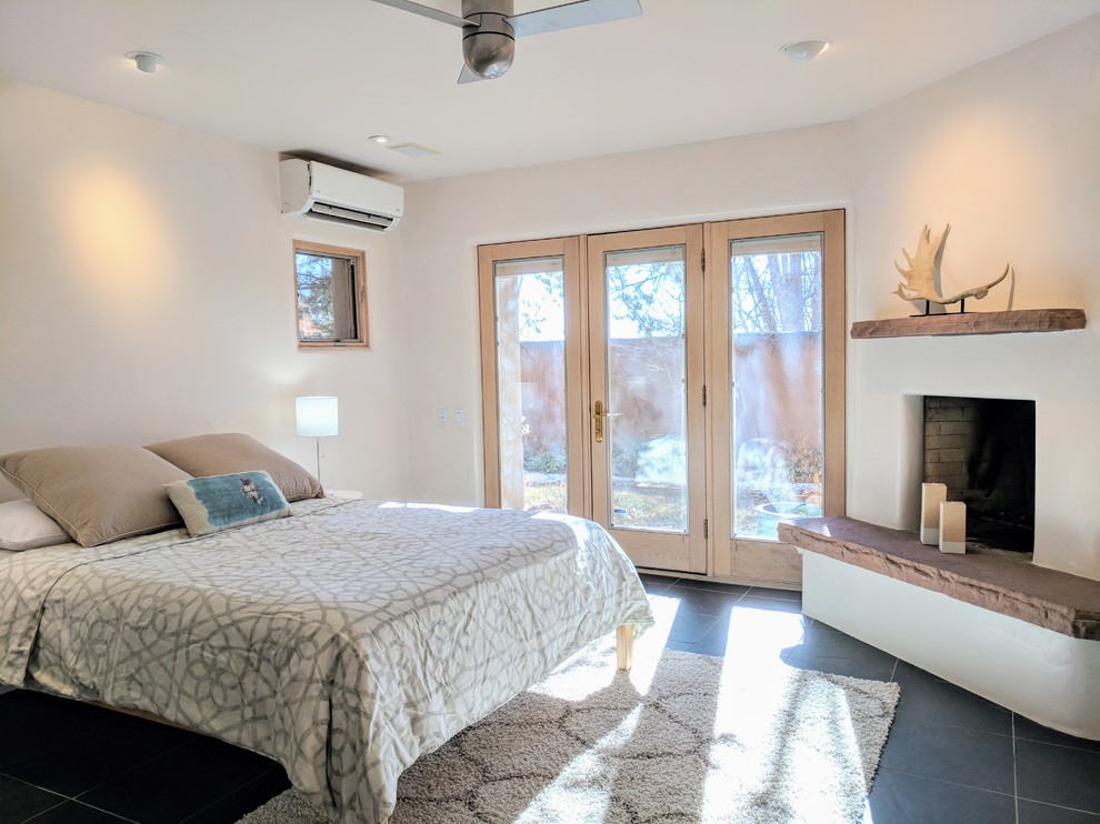 他の地域にある中くらいなサンタフェスタイルのおしゃれな主寝室 (白い壁、スレートの床、コーナー設置型暖炉、漆喰の暖炉まわり、黒い床) のインテリア