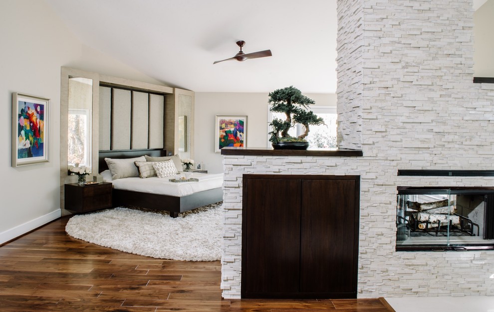 Cette photo montre une grande chambre parentale tendance avec un sol en bois brun, une cheminée double-face, un manteau de cheminée en pierre et un sol marron.