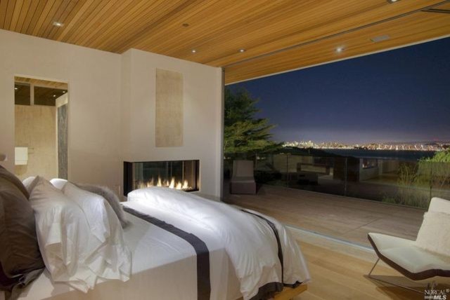 Expansive contemporary bedroom in San Francisco with beige walls, a two-sided fireplace, a stone fireplace surround and light hardwood flooring.