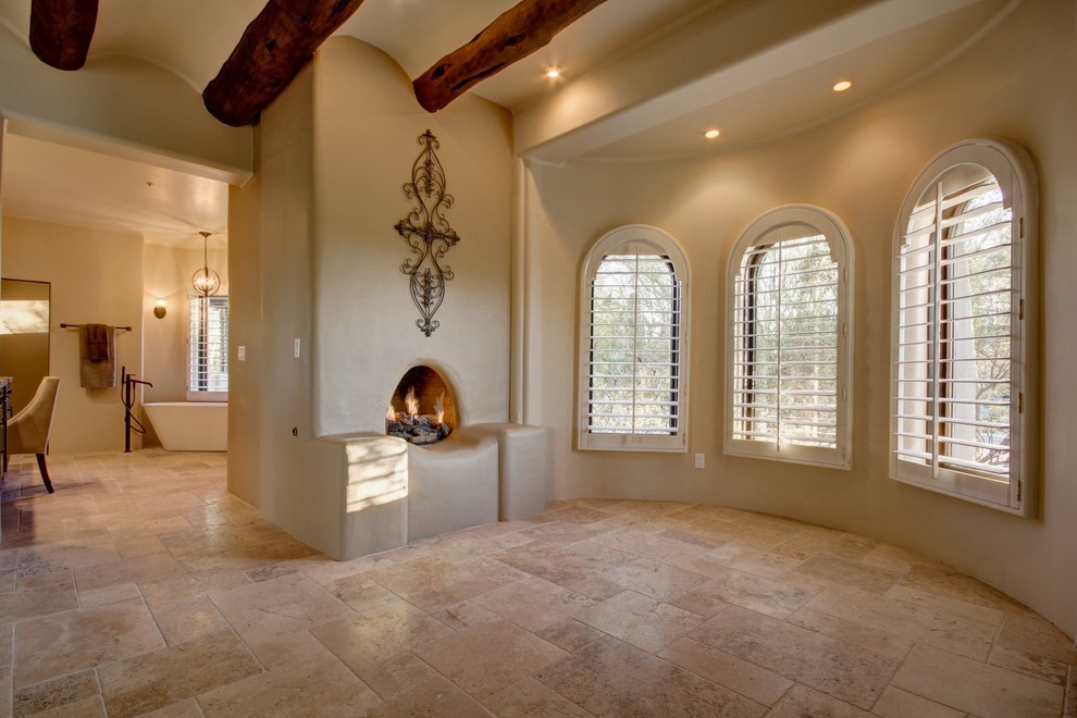 フェニックスにある巨大なサンタフェスタイルのおしゃれな主寝室 (ベージュの壁、トラバーチンの床、標準型暖炉、漆喰の暖炉まわり) のインテリア