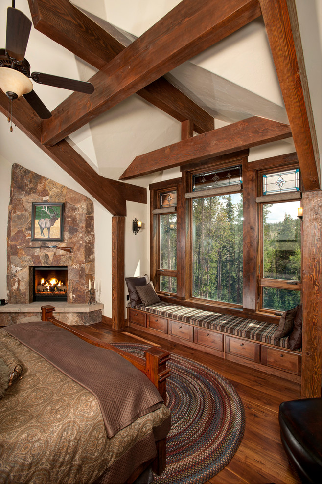 Diseño de dormitorio rústico con paredes beige, suelo de madera oscura, chimenea de esquina y marco de chimenea de piedra