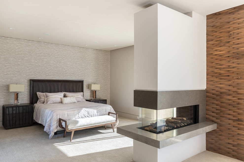 Diseño de dormitorio principal moderno grande con paredes blancas, moqueta, chimenea de doble cara, marco de chimenea de piedra y suelo beige
