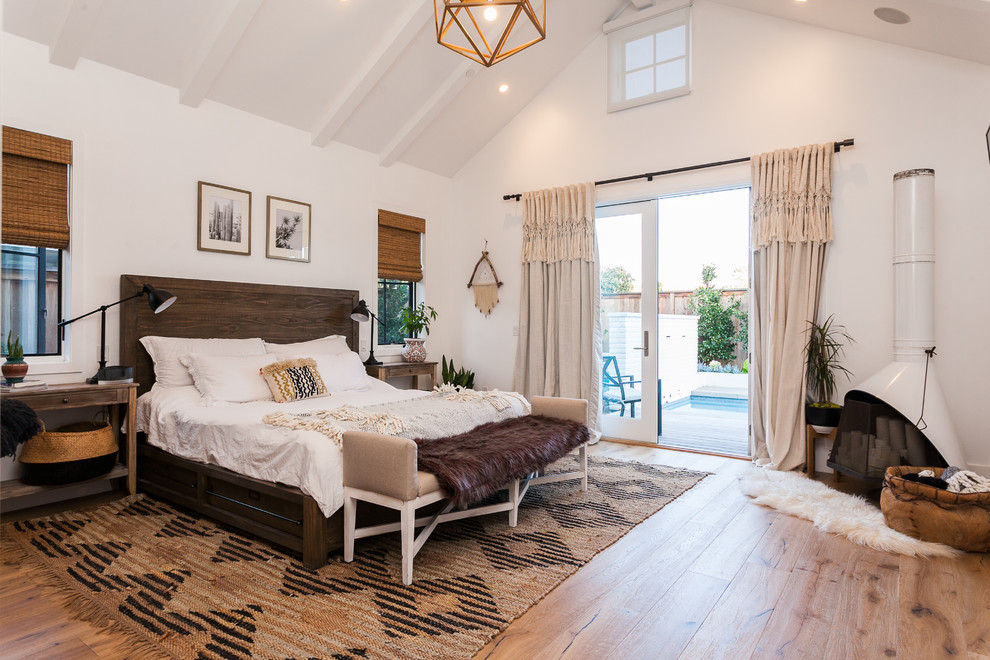 Diseño de dormitorio principal campestre con paredes blancas, suelo de madera en tonos medios, estufa de leña, marco de chimenea de metal y suelo beige