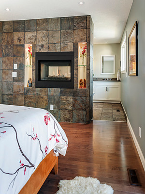 Modelo de dormitorio principal clásico renovado grande con paredes azules, suelo de madera en tonos medios, chimenea de doble cara y marco de chimenea de piedra