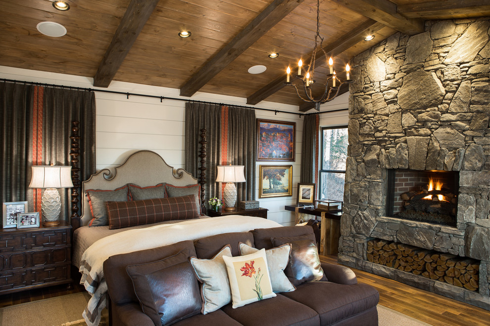 Réalisation d'une grande chambre parentale chalet avec un mur blanc, une cheminée standard, un manteau de cheminée en pierre et un sol en bois brun.