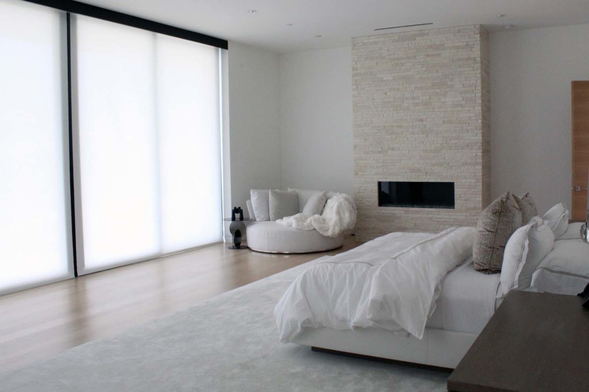 Modelo de dormitorio principal moderno grande con paredes blancas, suelo de madera clara, chimenea lineal, marco de chimenea de piedra y suelo beige