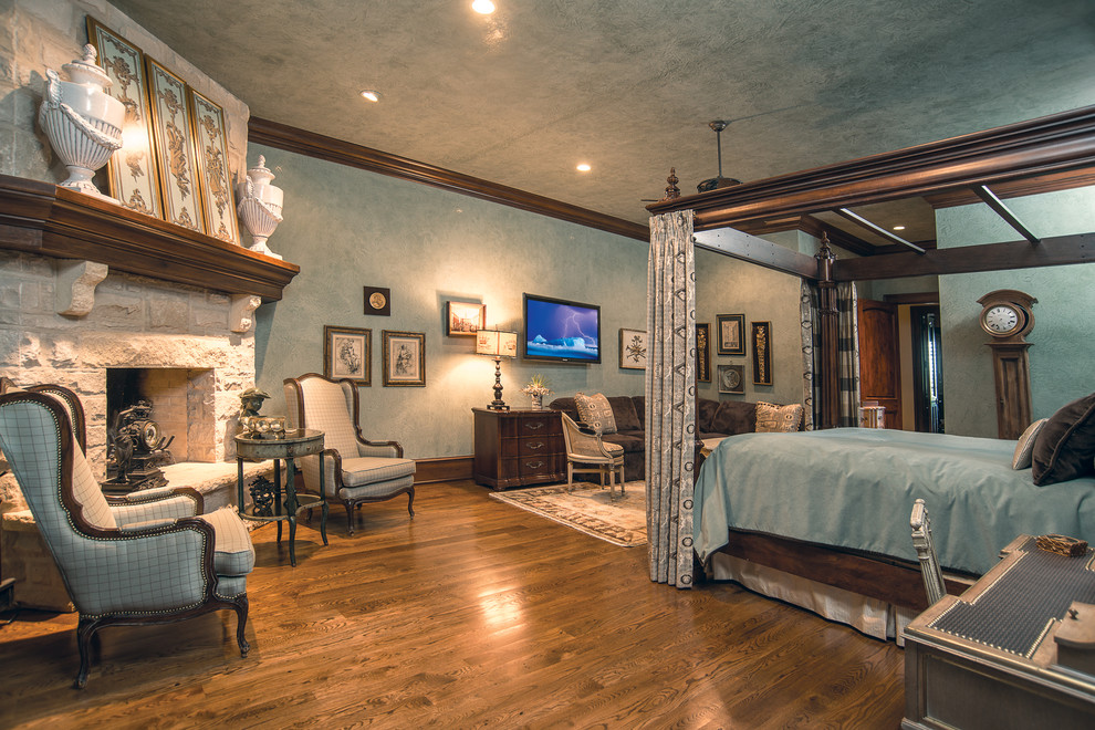 Ejemplo de dormitorio principal rural de tamaño medio con paredes azules, suelo de madera en tonos medios, todas las chimeneas y marco de chimenea de piedra