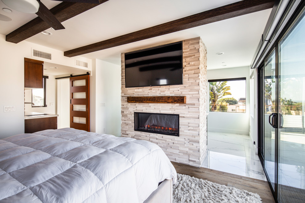 Modelo de dormitorio principal costero grande con paredes blancas, suelo laminado, chimenea de doble cara, marco de chimenea de baldosas y/o azulejos y suelo marrón