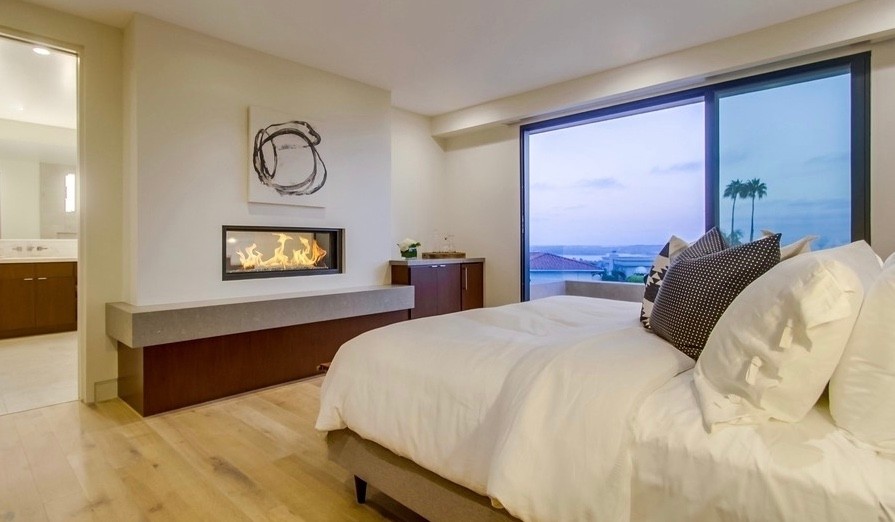 Imagen de dormitorio principal contemporáneo grande con paredes blancas, suelo de madera clara, chimenea de doble cara, marco de chimenea de yeso y suelo beige