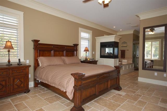 На фото: большая хозяйская спальня в классическом стиле с бежевыми стенами, полом из керамической плитки, двусторонним камином и фасадом камина из дерева