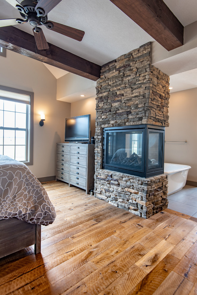 Ejemplo de dormitorio principal rural con paredes beige, suelo de madera clara, chimenea de doble cara, marco de chimenea de piedra y suelo marrón