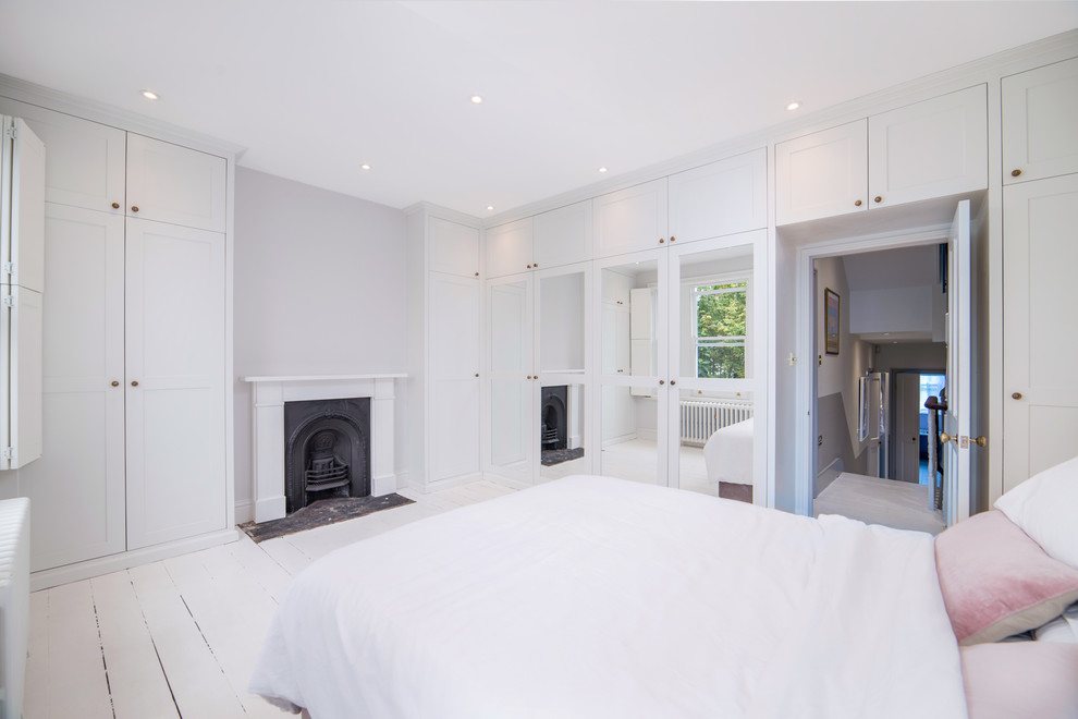 Klassisches Hauptschlafzimmer mit weißer Wandfarbe, gebeiztem Holzboden, Kamin, Kaminumrandung aus Holz und weißem Boden in London