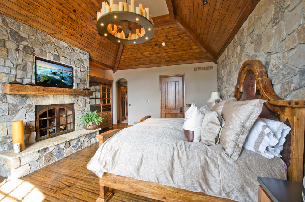 Cette image montre une grande chambre parentale chalet avec un mur gris, un sol en bois brun, une cheminée standard, un manteau de cheminée en pierre et un sol marron.