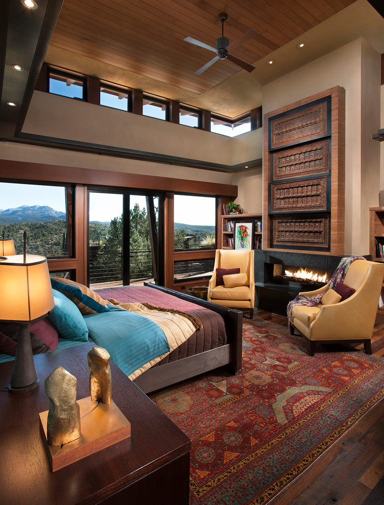 Modelo de dormitorio principal de estilo americano grande con paredes beige, suelo de madera en tonos medios, chimenea lineal, marco de chimenea de piedra y suelo marrón