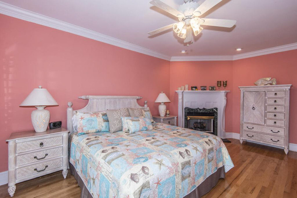 Modelo de dormitorio principal marinero grande con paredes rosas, suelo de madera en tonos medios, chimenea de esquina, marco de chimenea de piedra y suelo marrón