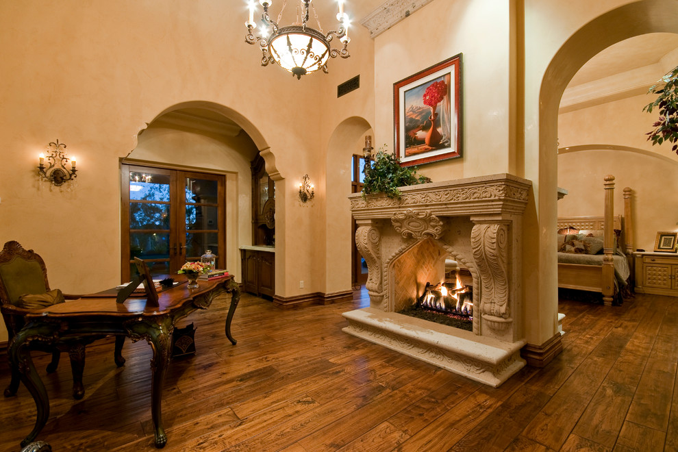 Photo of an expansive mediterranean master bedroom in Phoenix with beige walls, dark hardwood flooring, a two-sided fireplace, a stone fireplace surround and brown floors.