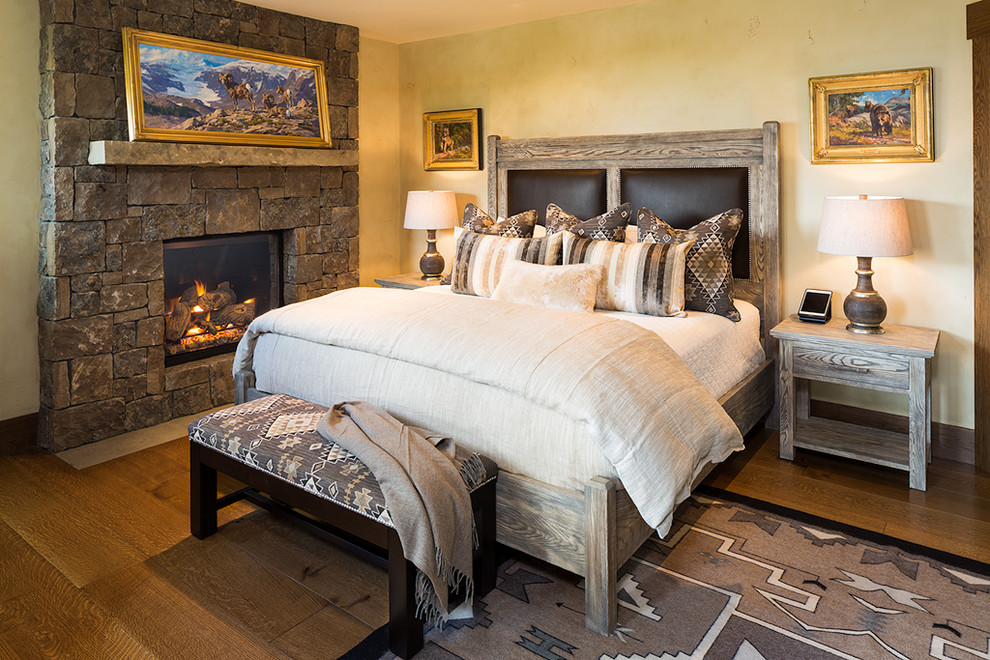 Cette image montre une grande chambre d'amis chalet avec un sol en bois brun, une cheminée standard, un manteau de cheminée en pierre, un mur beige et un sol marron.