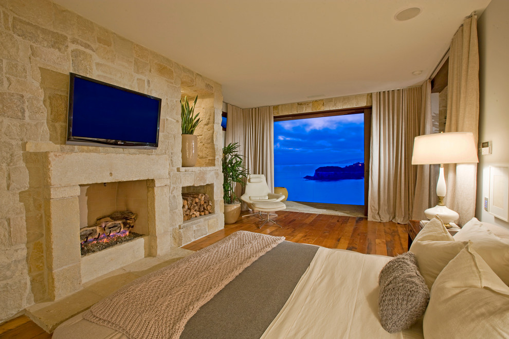 Mediterranes Schlafzimmer mit Kaminumrandung aus Stein, weißer Wandfarbe, braunem Holzboden und Kamin in Orange County