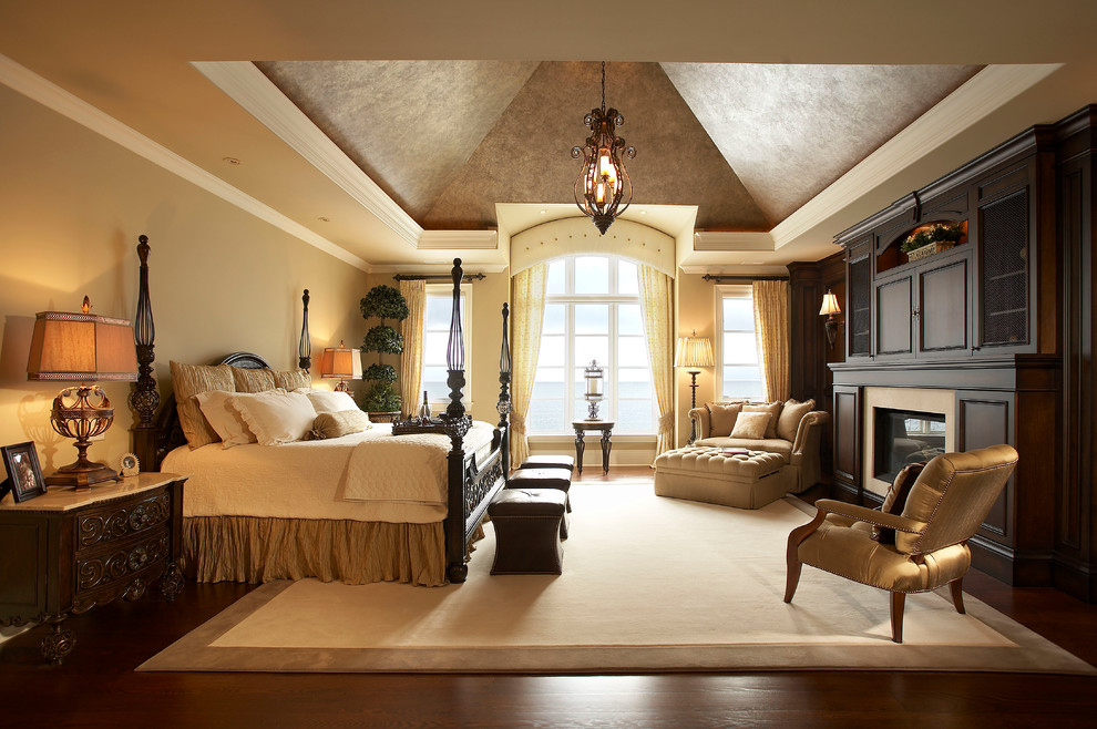 На фото: большая хозяйская спальня в классическом стиле с бежевыми стенами, темным паркетным полом, стандартным камином и фасадом камина из дерева с
