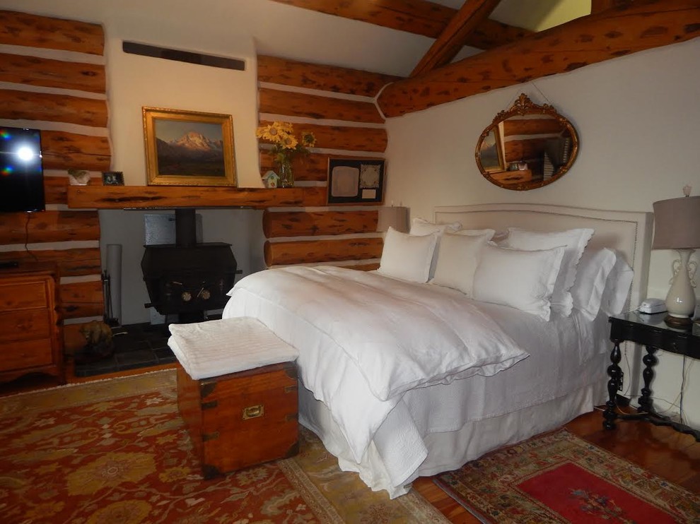 Ejemplo de dormitorio principal rústico de tamaño medio con paredes blancas, suelo de madera en tonos medios, estufa de leña, marco de chimenea de metal y suelo marrón