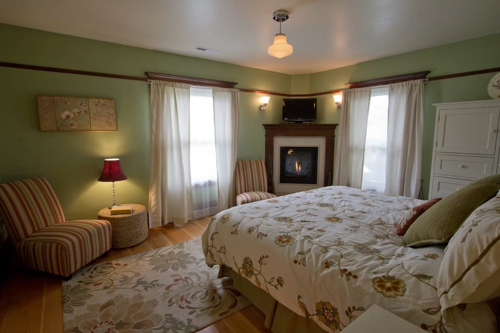 Modelo de dormitorio principal de estilo americano grande con paredes verdes, suelo de madera clara, chimenea de esquina y marco de chimenea de madera