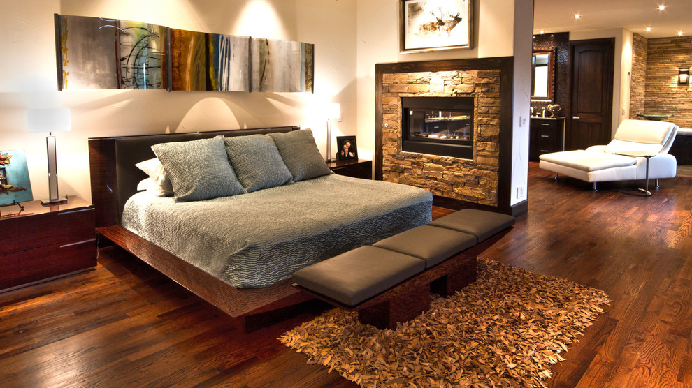 Ejemplo de dormitorio principal minimalista grande con paredes beige, suelo de madera oscura, chimenea de doble cara y marco de chimenea de piedra