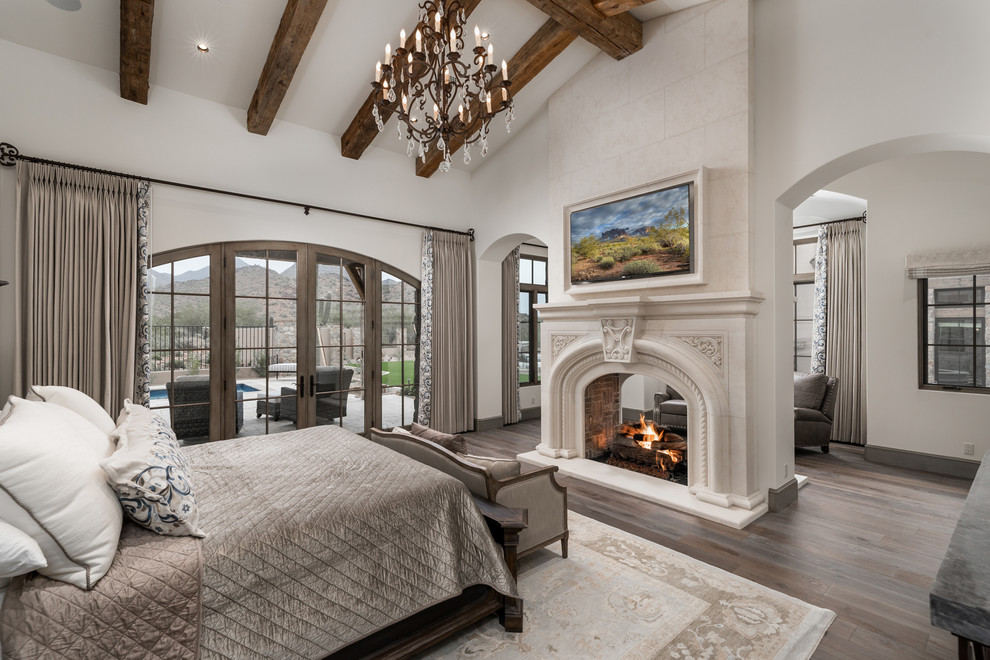 Expansive rustic master bedroom in Phoenix with white walls, medium hardwood flooring, a two-sided fireplace, a stone fireplace surround, brown floors and exposed beams.