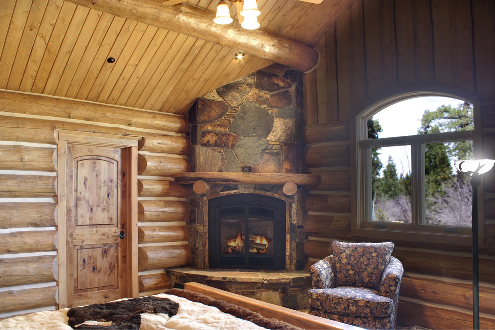 Modelo de dormitorio principal rural de tamaño medio con paredes marrones, suelo de madera en tonos medios, chimenea de esquina, marco de chimenea de piedra y suelo marrón