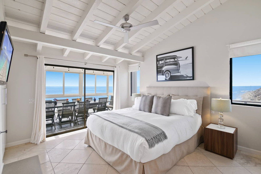 ロサンゼルスにある中くらいなビーチスタイルのおしゃれな主寝室 (グレーの壁、ライムストーンの床、標準型暖炉、漆喰の暖炉まわり、ベージュの床) のインテリア