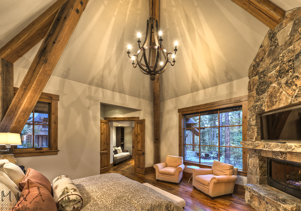 Modelo de habitación de invitados de estilo americano con paredes beige, suelo de madera oscura, chimenea de esquina, marco de chimenea de piedra y suelo marrón