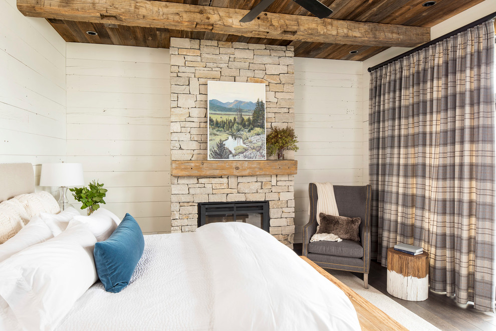 Modelo de dormitorio principal rural de tamaño medio con paredes blancas, suelo de madera oscura, todas las chimeneas, marco de chimenea de piedra y suelo marrón