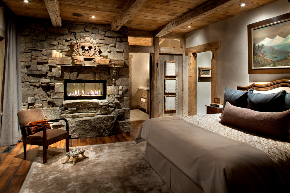 Modelo de dormitorio rural con marco de chimenea de piedra, chimenea de doble cara y suelo de madera en tonos medios
