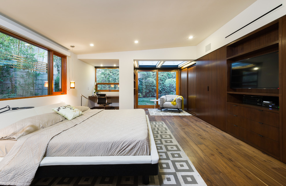 Diseño de dormitorio principal y televisión actual de tamaño medio sin chimenea con paredes blancas, suelo de madera en tonos medios y suelo marrón