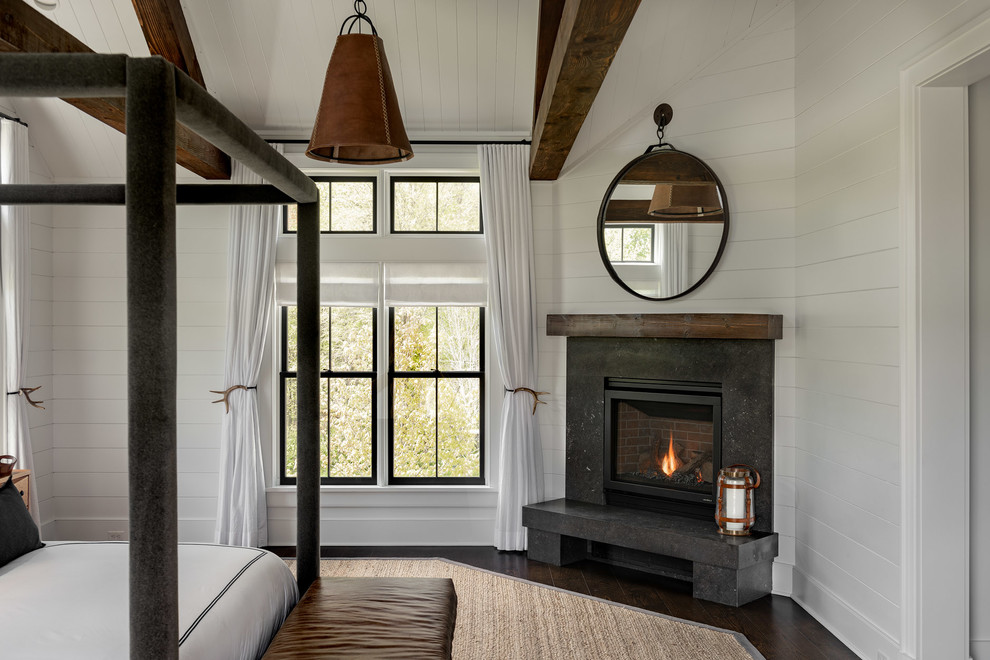 Diseño de dormitorio principal campestre grande con paredes blancas, suelo de madera oscura, chimenea de esquina, marco de chimenea de piedra y suelo marrón
